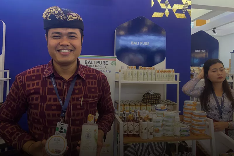 Seorang pria tersenyum di stand pameran Bali Pure Home Industry, memegang produk VCO dan sabun alami. Terdapat rak penuh produk kecantikan dan perawatan